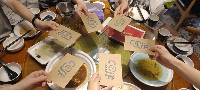 A group of people are holding up cards with the word "COU" on them, sitting around a table with various dishes and bowls.  (Captioned by AI)