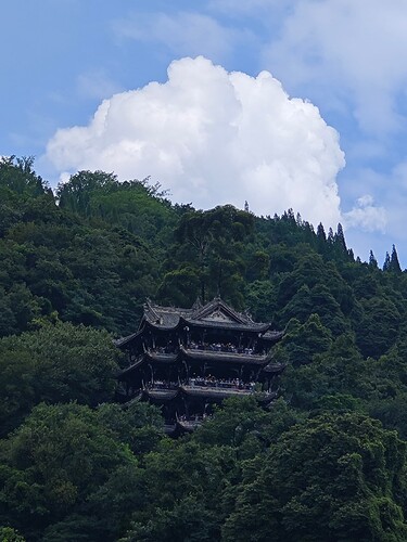 The image depicts a majestic, multi-tiered pagoda-style building nestled among lush green trees, with a large white cloud looming in the background against a blue sky. (由 AI 生成标题)