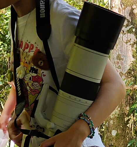The image captures a person, visible from the torso up, holding a large camera lens, with a strap and a colorful shirt. The person is wearing a white t-shirt with a cartoon design and the word "HAPPY" in red letters. A black camera strap is slung over their shoulder, and... (由 AI 生成标题)