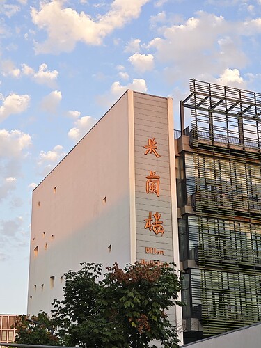 The image shows a tall, white building with orange Chinese characters on the side and a glass section on the right, set against a blue sky with fluffy clouds. (由 AI 生成标题)