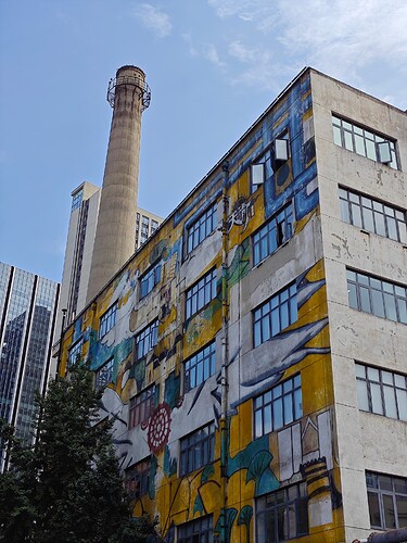 The image shows a large, multi-story building with a vibrant mural on its side, featuring a mix of yellow, blue, white, and green colors, set against a backdrop of a clear blue sky and a tall industrial chimney. (由 AI 生成标题)