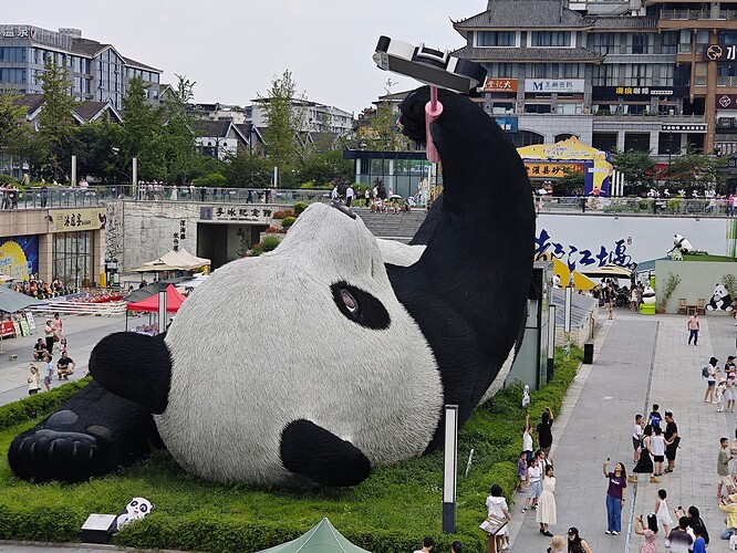The image depicts a large panda sculpture lying on its back with one leg raised and people walking around it. (由 AI 生成标题)