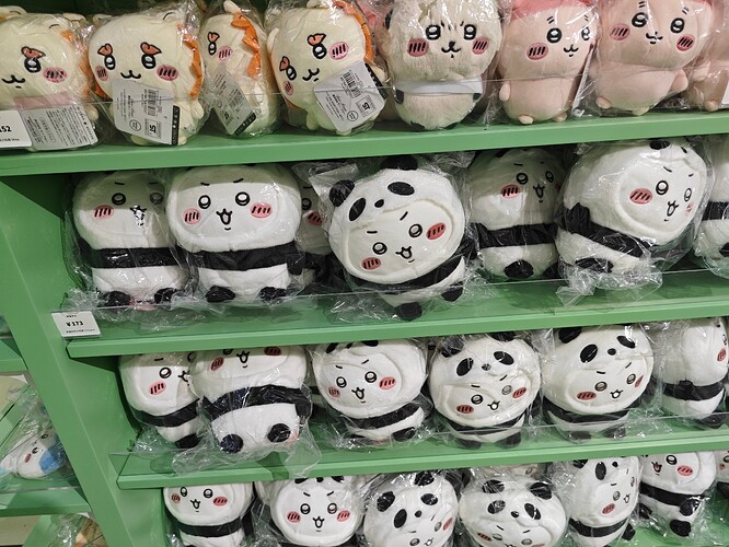 The image shows three green shelves stacked with plush toys individually wrapped in plastic and featuring cute, stylized faces. (由 AI 生成标题)