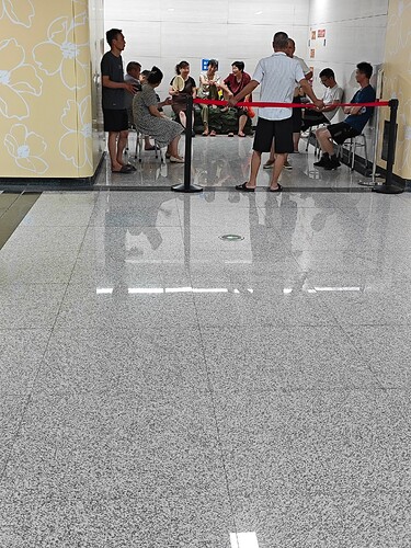 The image shows a group of people sitting and waiting in line, separated by a red rope. (由 AI 生成标题)